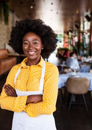 Portrait of positive African american young woman working professional confectioner in own coffee shop, looking at camera with toothy smile.